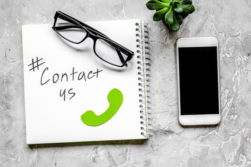 A note pad with Contact Us written on it and a mobile phone and pair of glasses on a desk