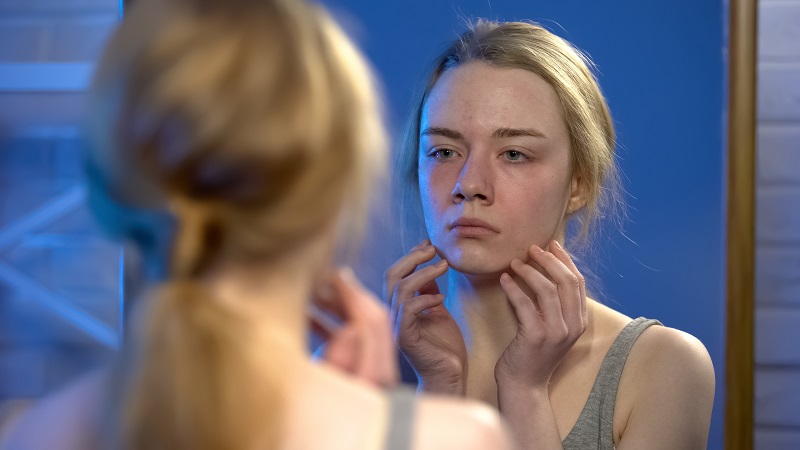 A female young adult looking at her face in the mirror. She looks concerned.