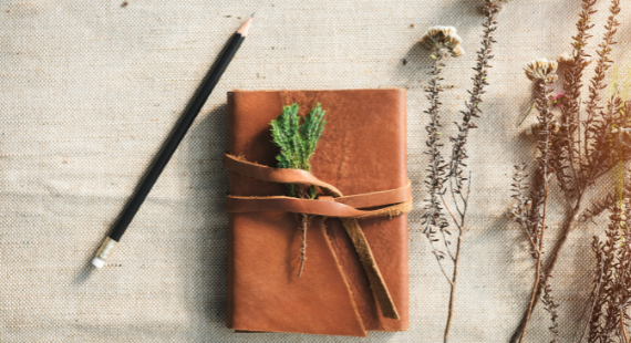 This image shows a leather-bound journal on a hessian cloth, with a pencil and some dried flowers beside it.