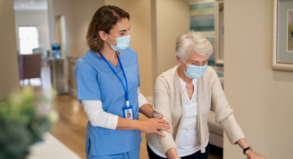 This image shows a member of staff in a nursing home helping an older lady who is using a walking frame, with both wearing masks for infection control.