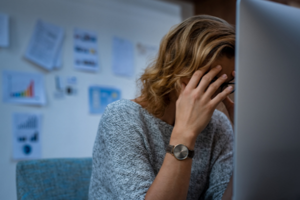 A woman holds her head in her hands in stress as she sits in front of a computer screen in a workplace office setting.