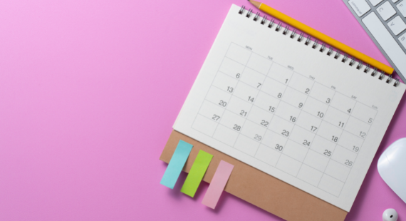 A calendar sits on a desk beside a computer keyboard, with a pencil and post-it notes ready to mark important dates.