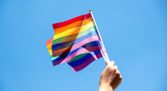 A person waves a Pride rainbow flag in the air against the backdrop of a bright blue sky