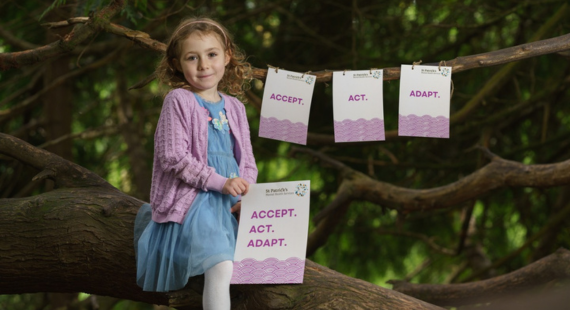 Photo of a young girl sitting on the branch of a tree, holding a sign saying "Accept Act Adapt" with tags hanging on other tree branches with the same wording.