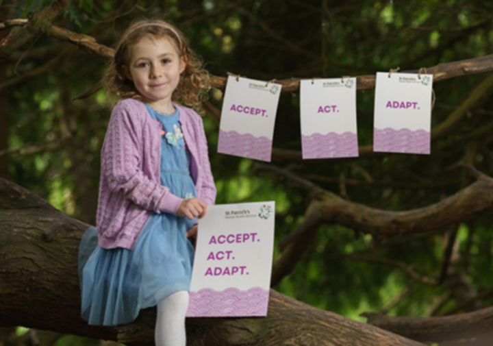 Photo of a young girl sitting on the branch of a tree, holding a sign saying "Accept Act Adapt" with tags hanging on other tree branches with the same wording.